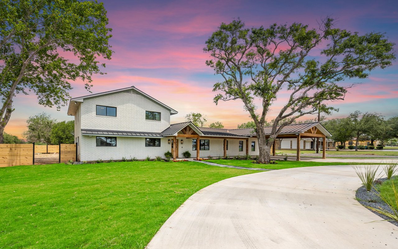 White-brick home with covered porch and carport beneath a colorful sunset sky.