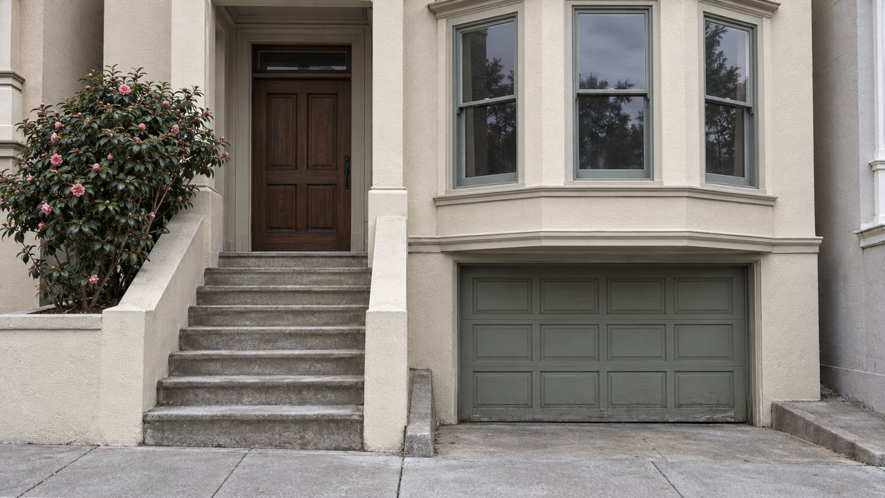 Cream stucco house with stacked gray-green bay windows, a recessed wood entrance, worn concrete steps and a single camellia beside the stair.