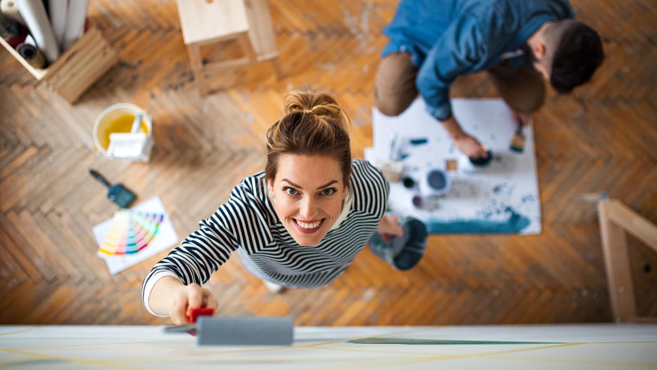 A top down view of a couple painting an interior wall with bright neutral colours, with a smiling woman looking up while using a paint roller, representing the low cost high impact cosmetic updates that maximize property appeal and yield the best home improvements ROI for resale.