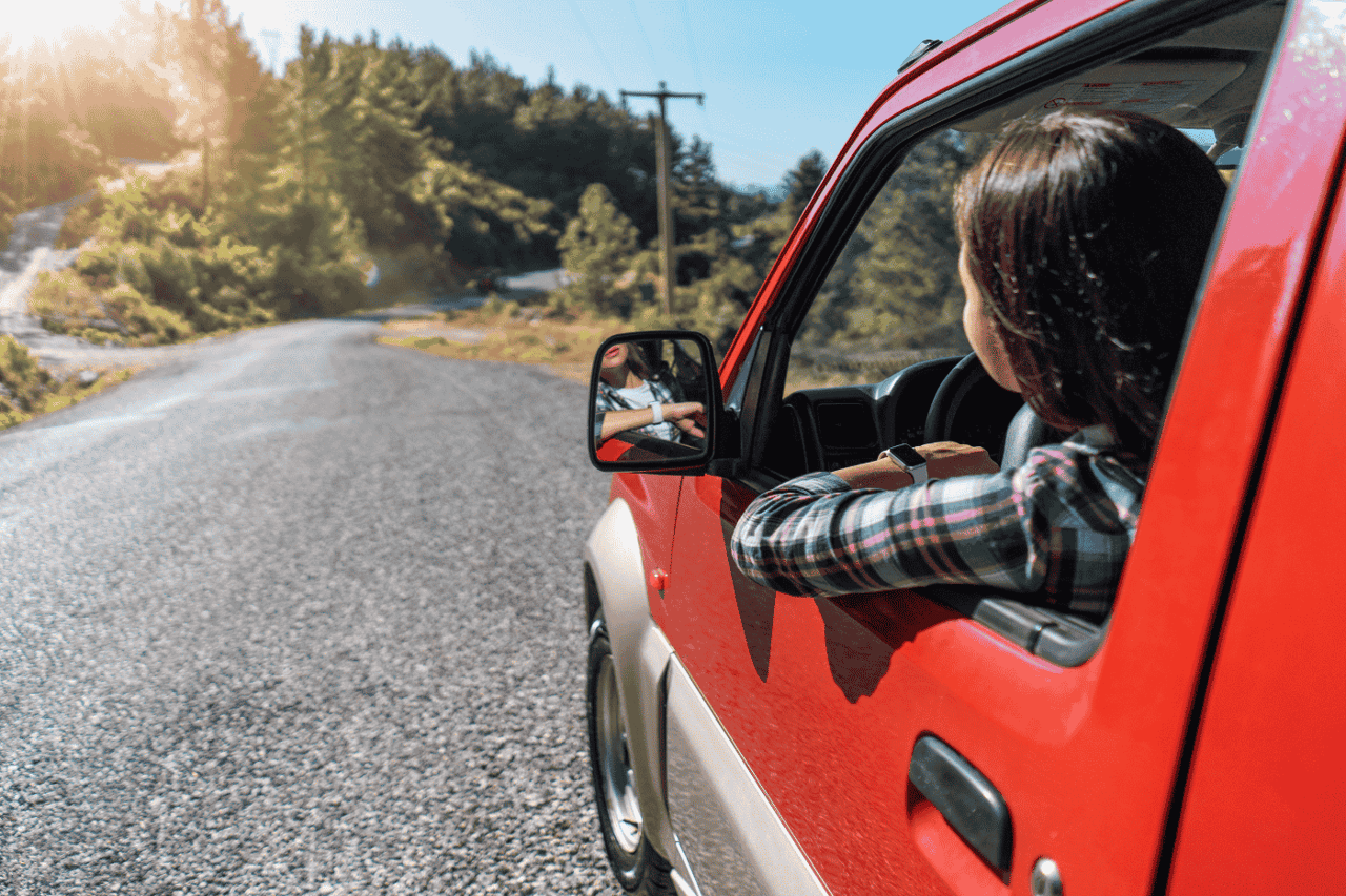 Woman driving red car along a sunny rural road with trees.