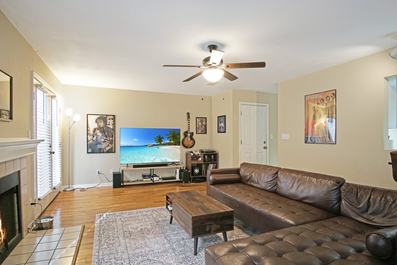 Living room at 529 Collinsford Road, Tallahassee, Florida, featuring a brown sectional sofa, patterned area rug, and wooden coffee table. The space includes hardwood floors, a ceiling fan with light fixture, a fireplace with tile surround, and a large TV setup with wall art and a guitar display, creating a warm and inviting entertainment area.