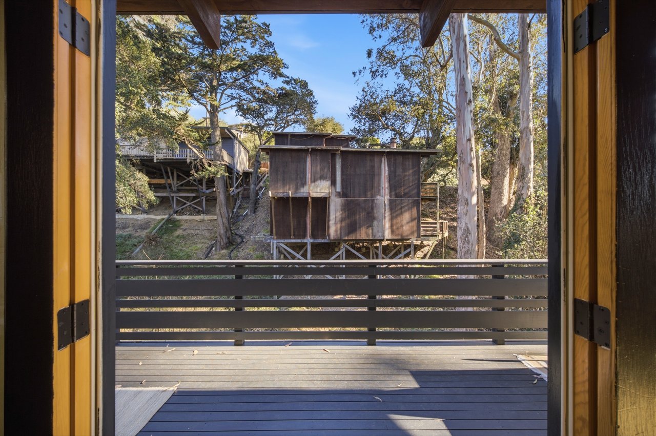 Wooden deck overlooking a wooded hillside with elevated structures, framed by open doors.