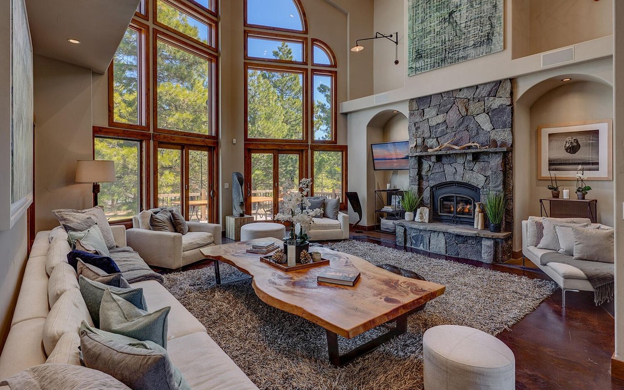 Furnished living room with tall windows overlooking trees and a floor-to-ceiling stone fireplace.