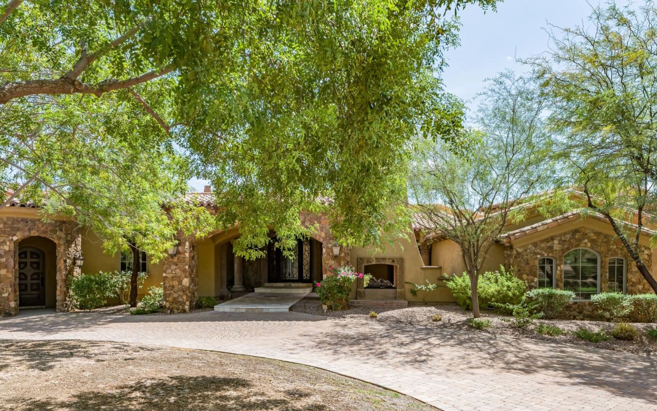 Stone-and-stucco home with arched entrances, a tiled roof, and a tree-shaded paved driveway.