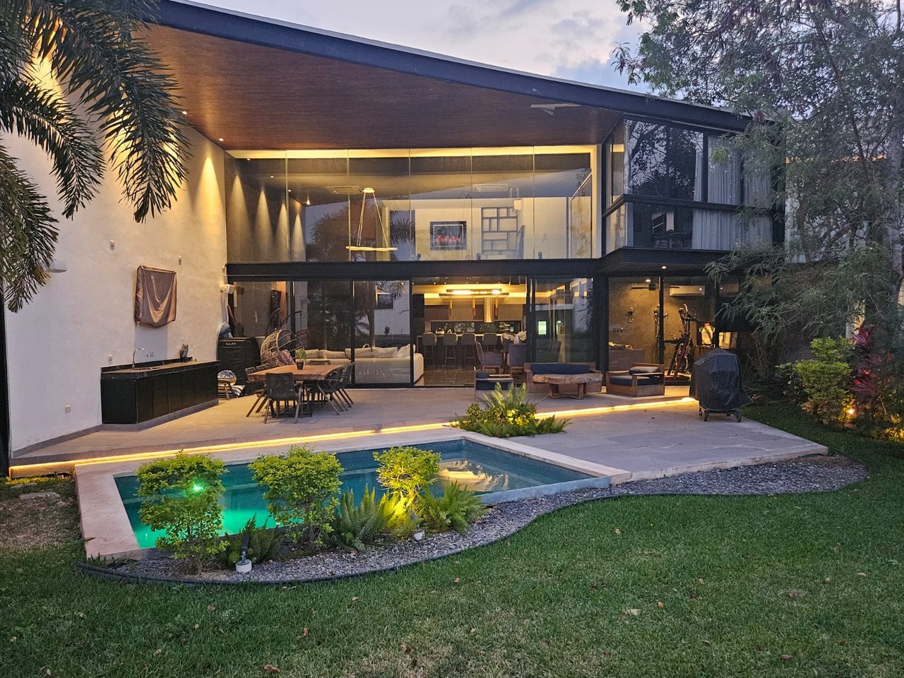 Modern glass-fronted home with a covered patio and illuminated pool at dusk.