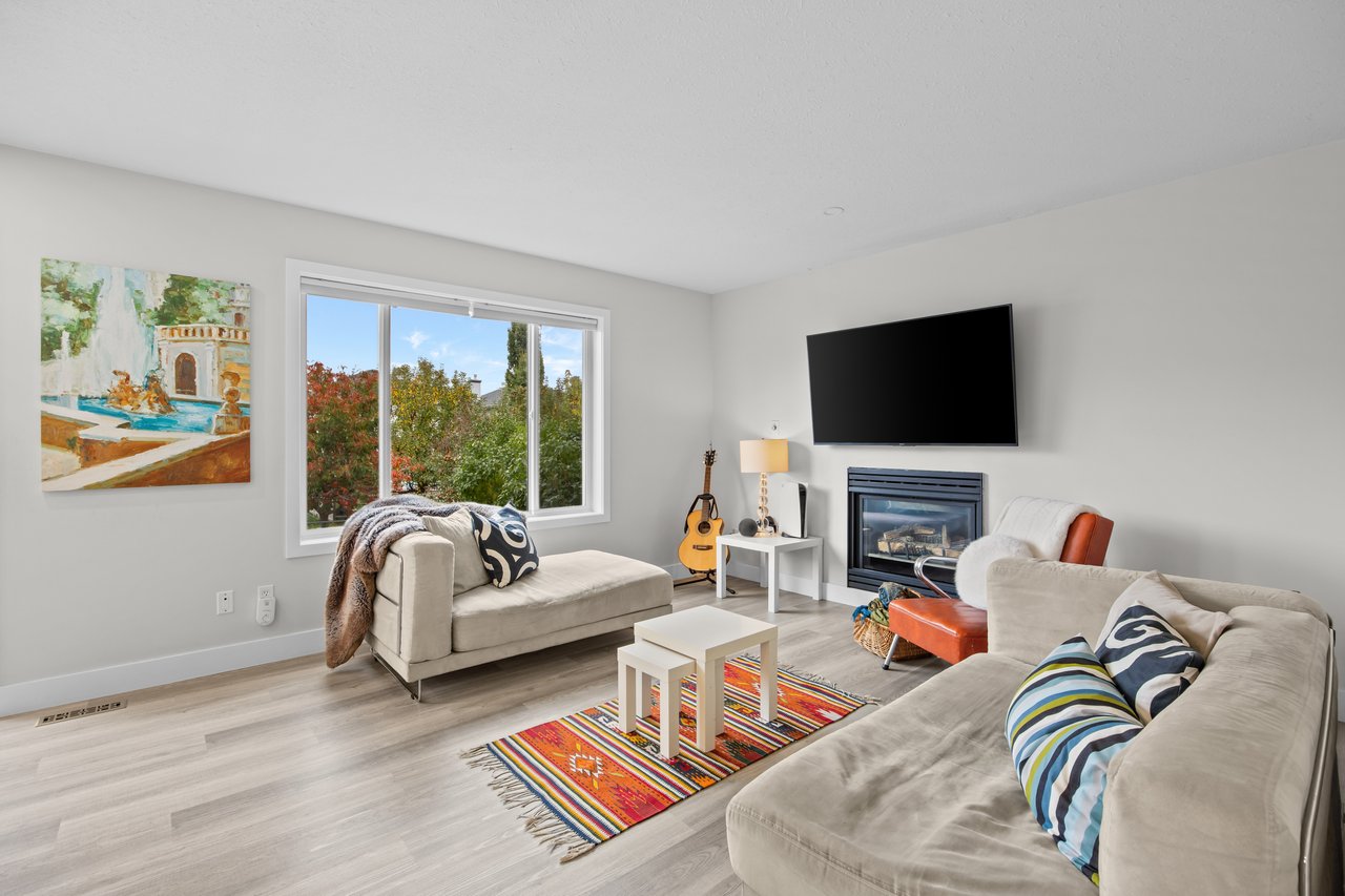 Living room with light sofas, a colorful rug, fireplace, and autumn foliage visible through the window.