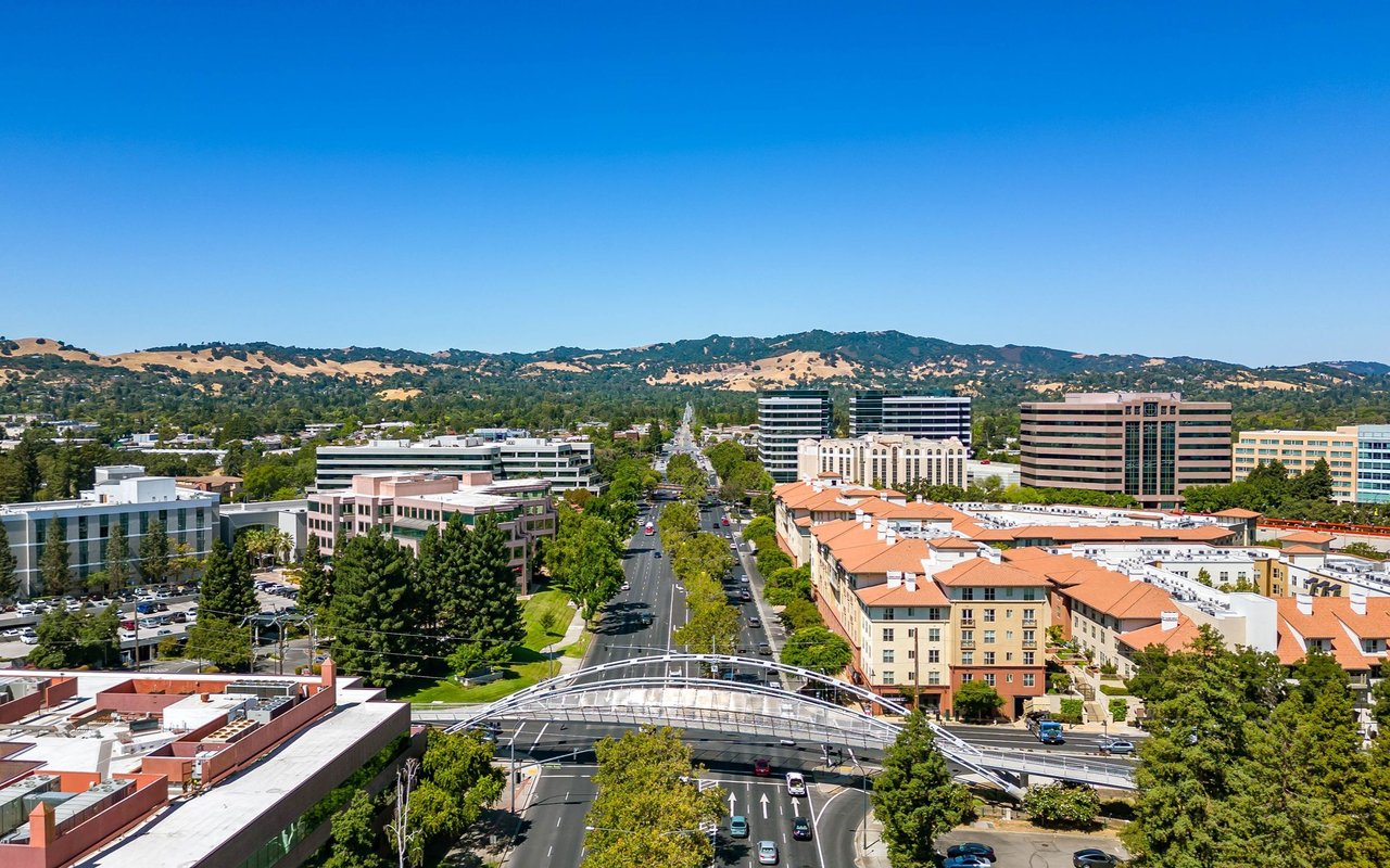 Aerial view of a business district with a tree-lined road, pedestrian bridge, and rolling hills.