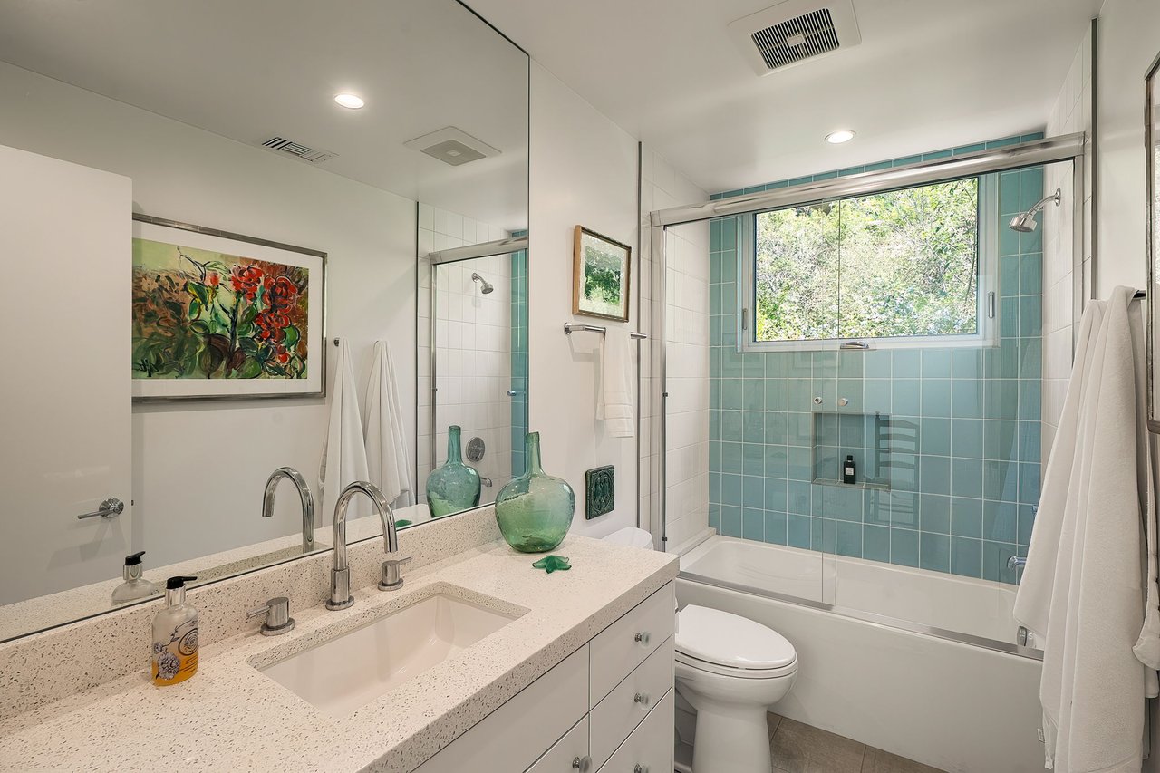 Bathroom with a speckled double-sink vanity and turquoise-tiled tub-shower beneath a wide window.