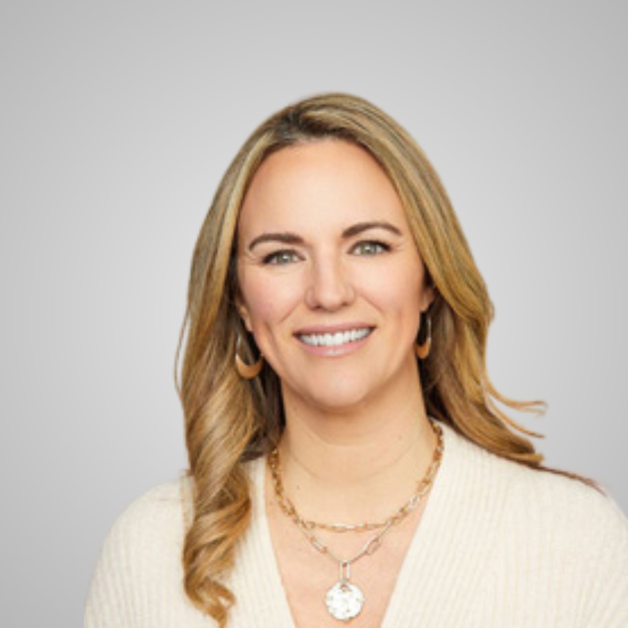 Smiling blonde woman in a white sweater with layered gold necklaces.