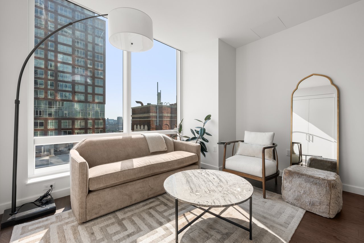 Furnished living room with a beige sofa and large windows overlooking nearby buildings.