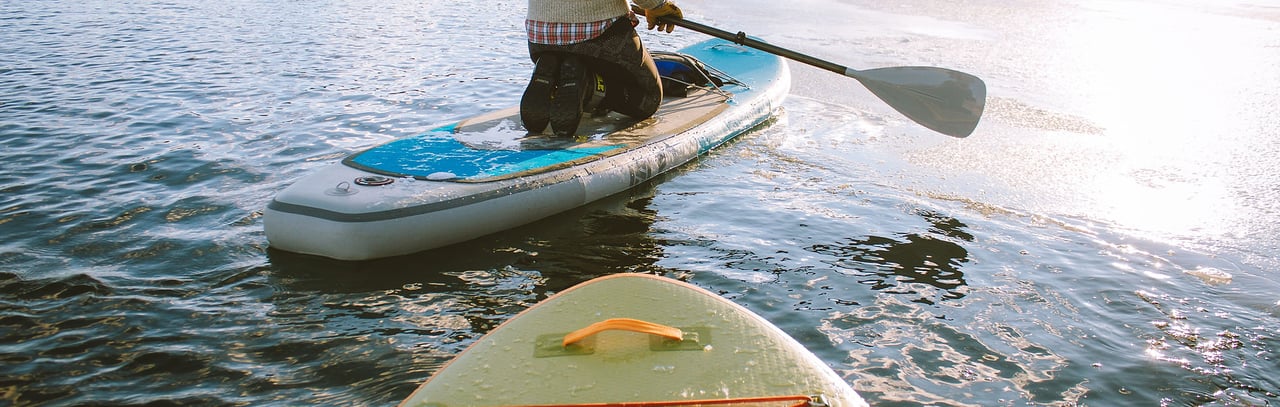 A paddleboarder kneels on a board on a sunlit lake beside another board.