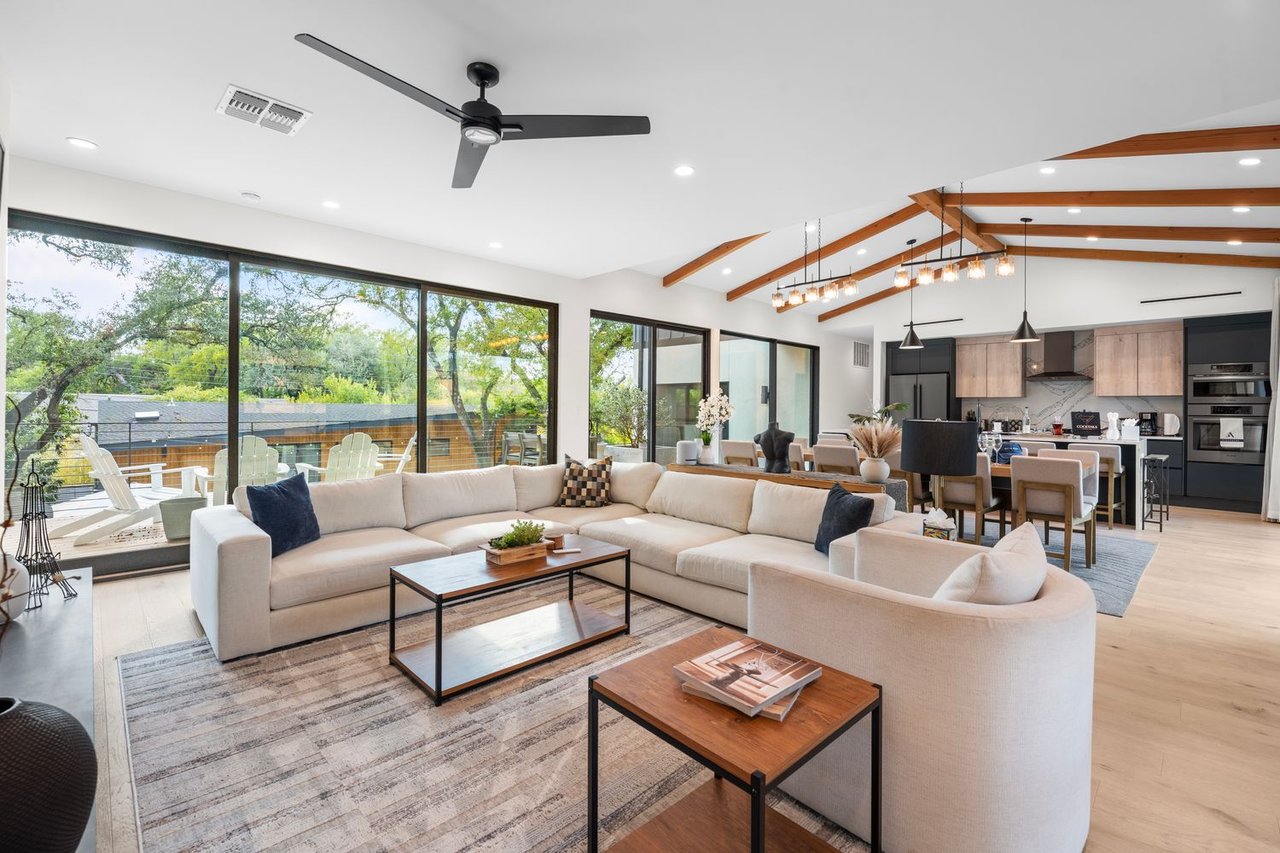 Open-plan living room with a cream sectional, exposed wood beams, and broad windows overlooking trees.
