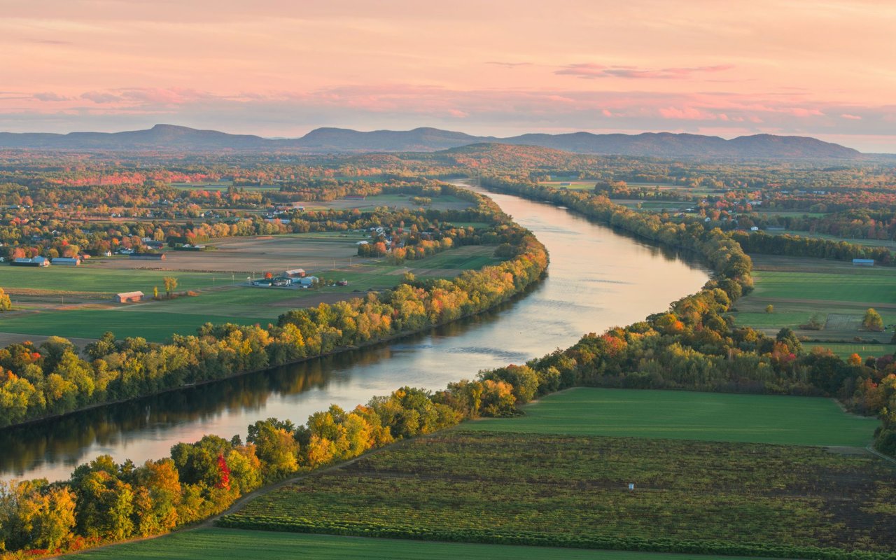 Winding river through autumn-colored farmland and wooded countryside, with distant hills under a pink sky.