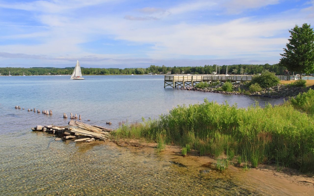 Suttons Bay shoreline with a sailboat, wooden pier, and clear shallow water.