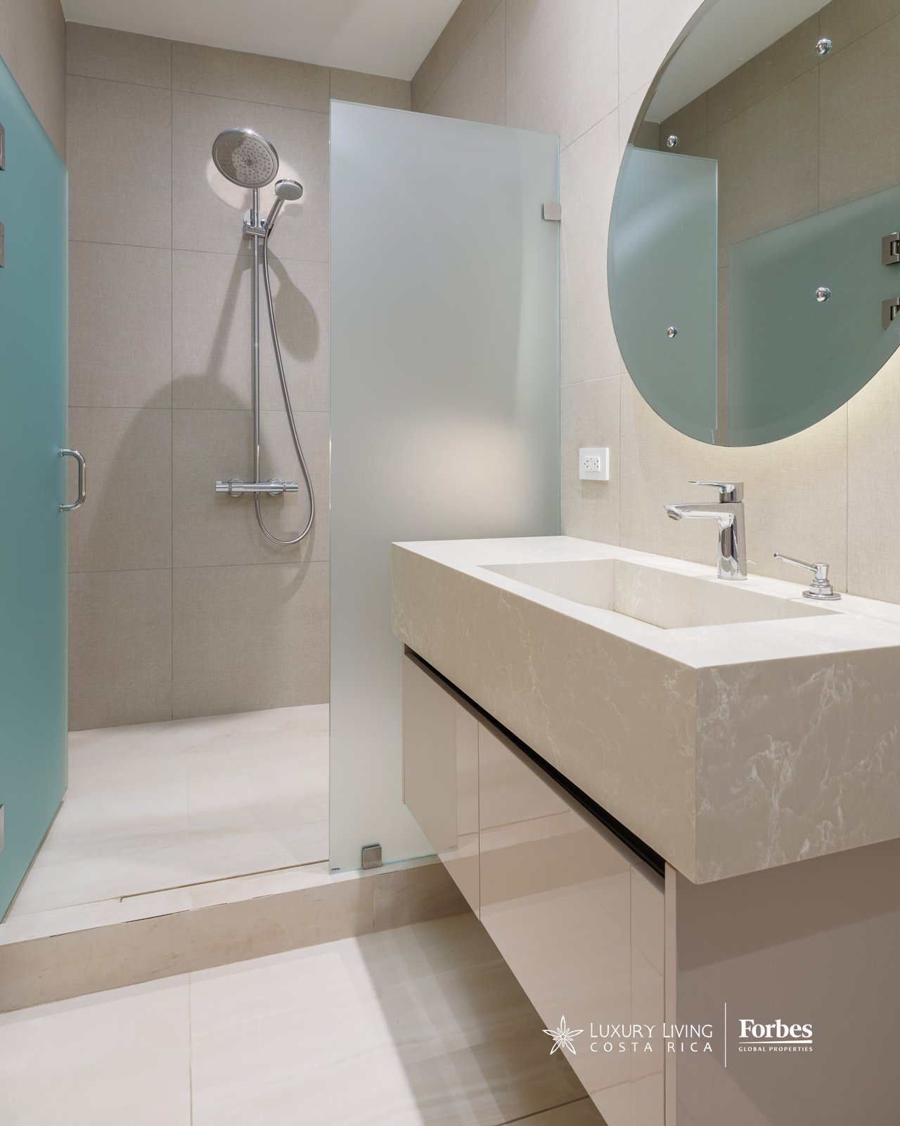 Modern bathroom with a stone vanity, round mirror, and glass-enclosed walk-in shower.