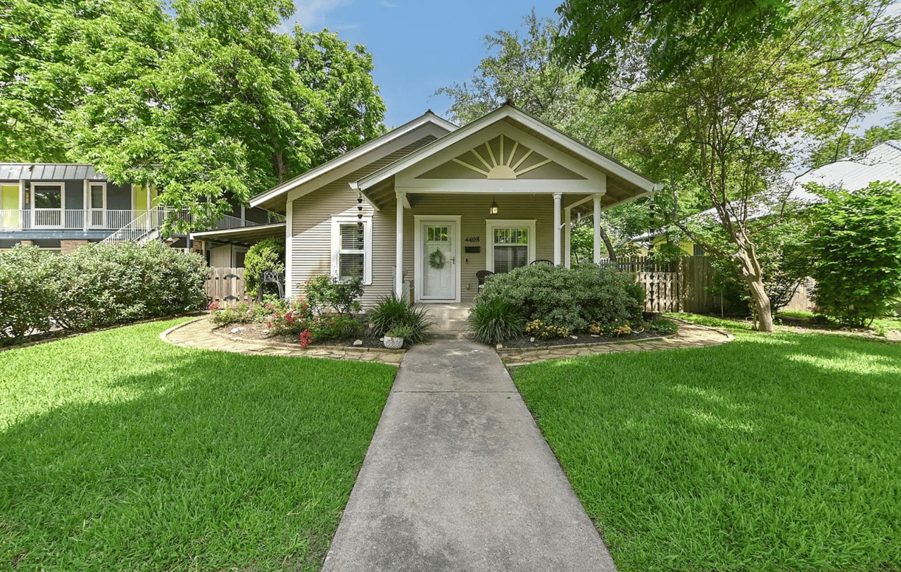 Beige house with a covered porch, sunburst gable, flower beds, and a walkway through the lawn.