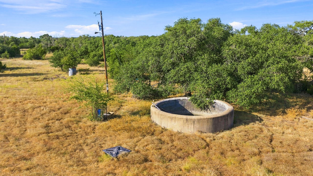 Ranch acreage with a circular concrete water trough among oak trees and dry grass.