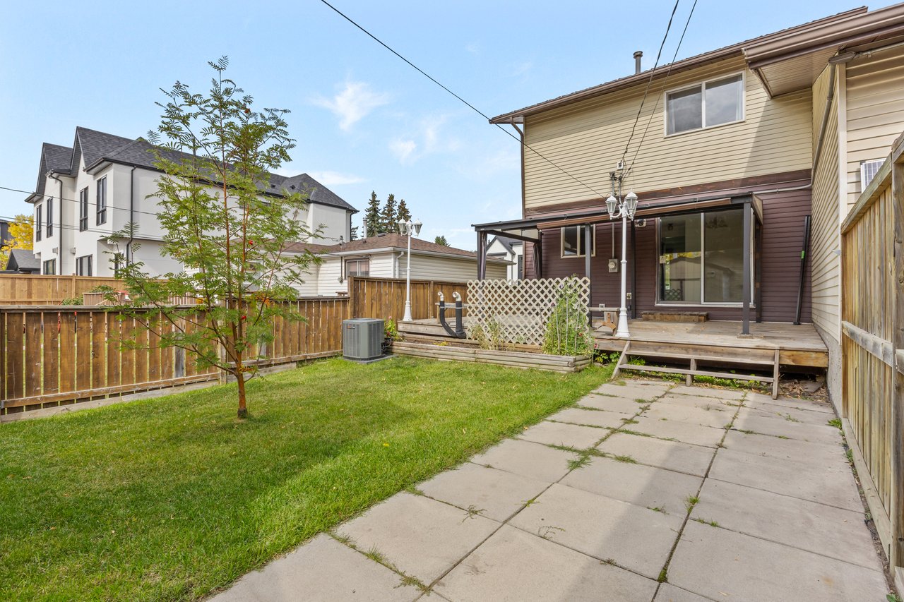 Fenced backyard with a concrete patio, green lawn, young tree, and raised wooden deck.