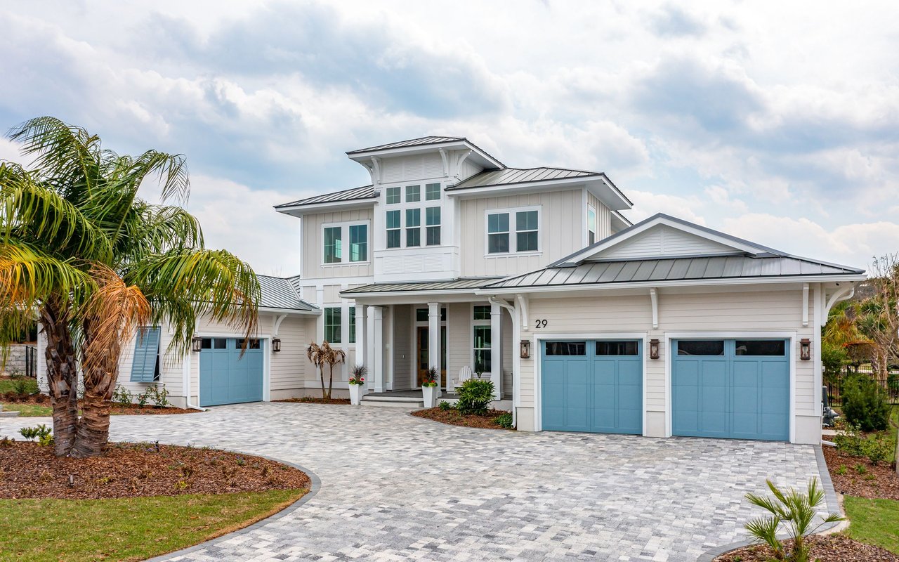 Modern two-story home with blue garage doors, metal roof, and palm-lined driveway.