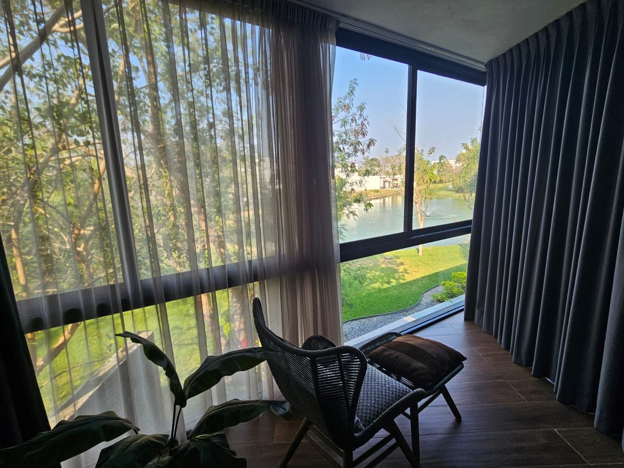 Furnished window seating area overlooking a lake, with dark curtains and a woven lounge chair.
