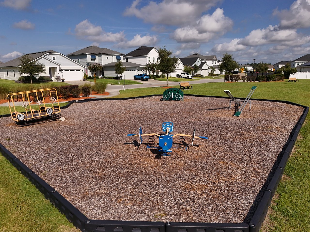 Neighborhood playground with wood-chip ground and play equipment, with homes along the street behind it.