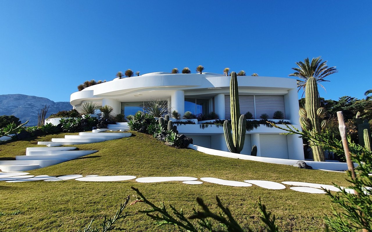 Modern white home on a landscaped hillside with cacti, palms, and distant mountains.