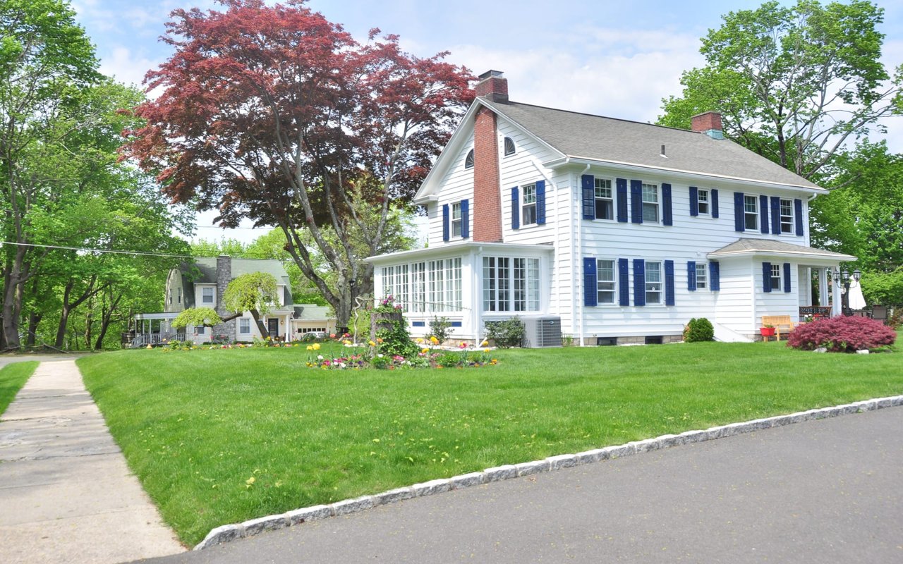 A white house with blue shutters with a red tree in the background and a green bush in front of the house.