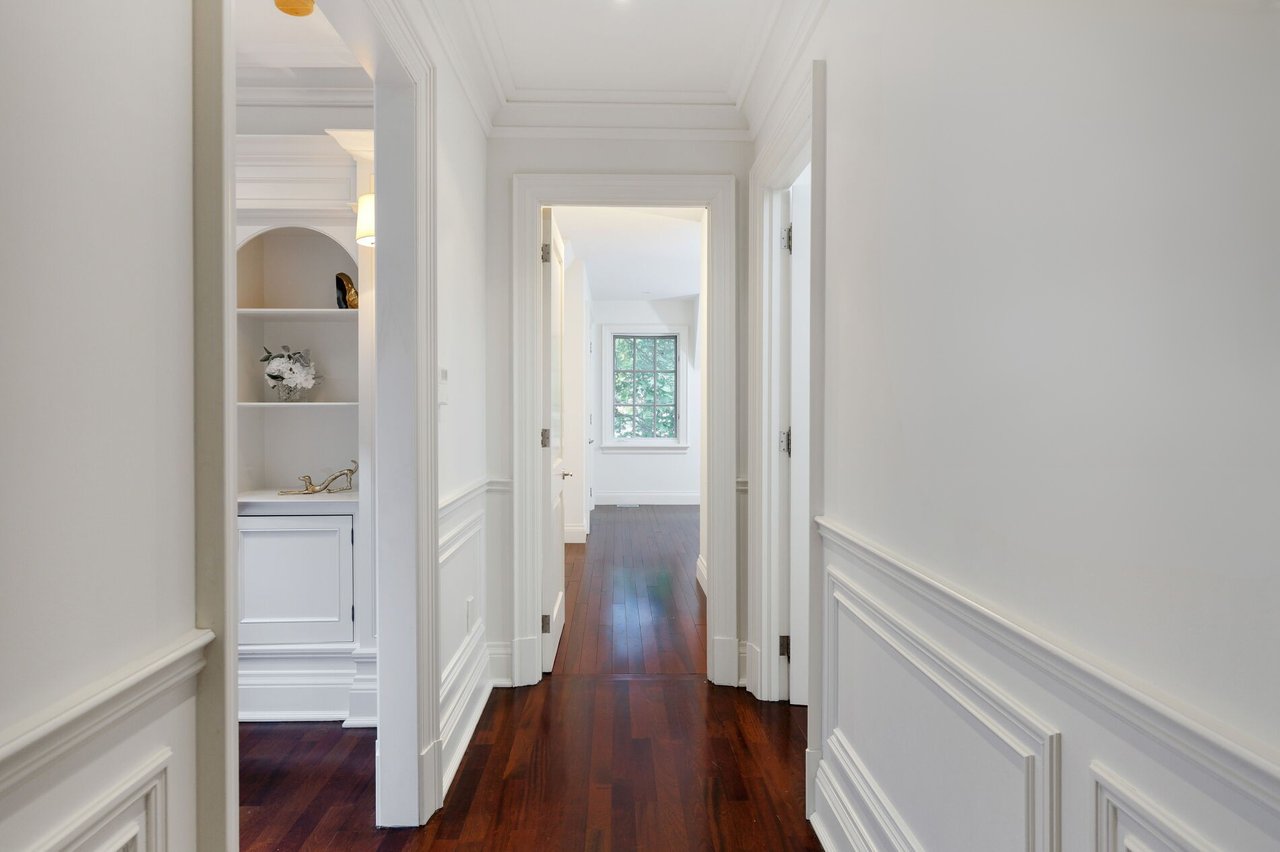 Bright hallway with dark hardwood floors, white paneled walls, built-in shelves and a window at the end.