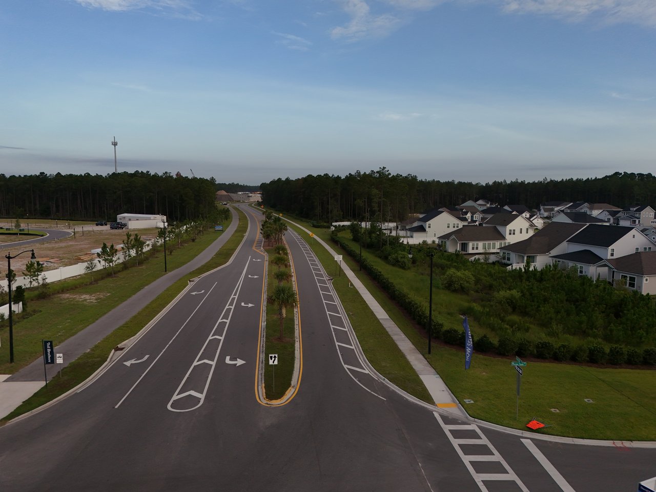 Residential development with a divided entrance road, new homes, pine trees, and open construction land.
