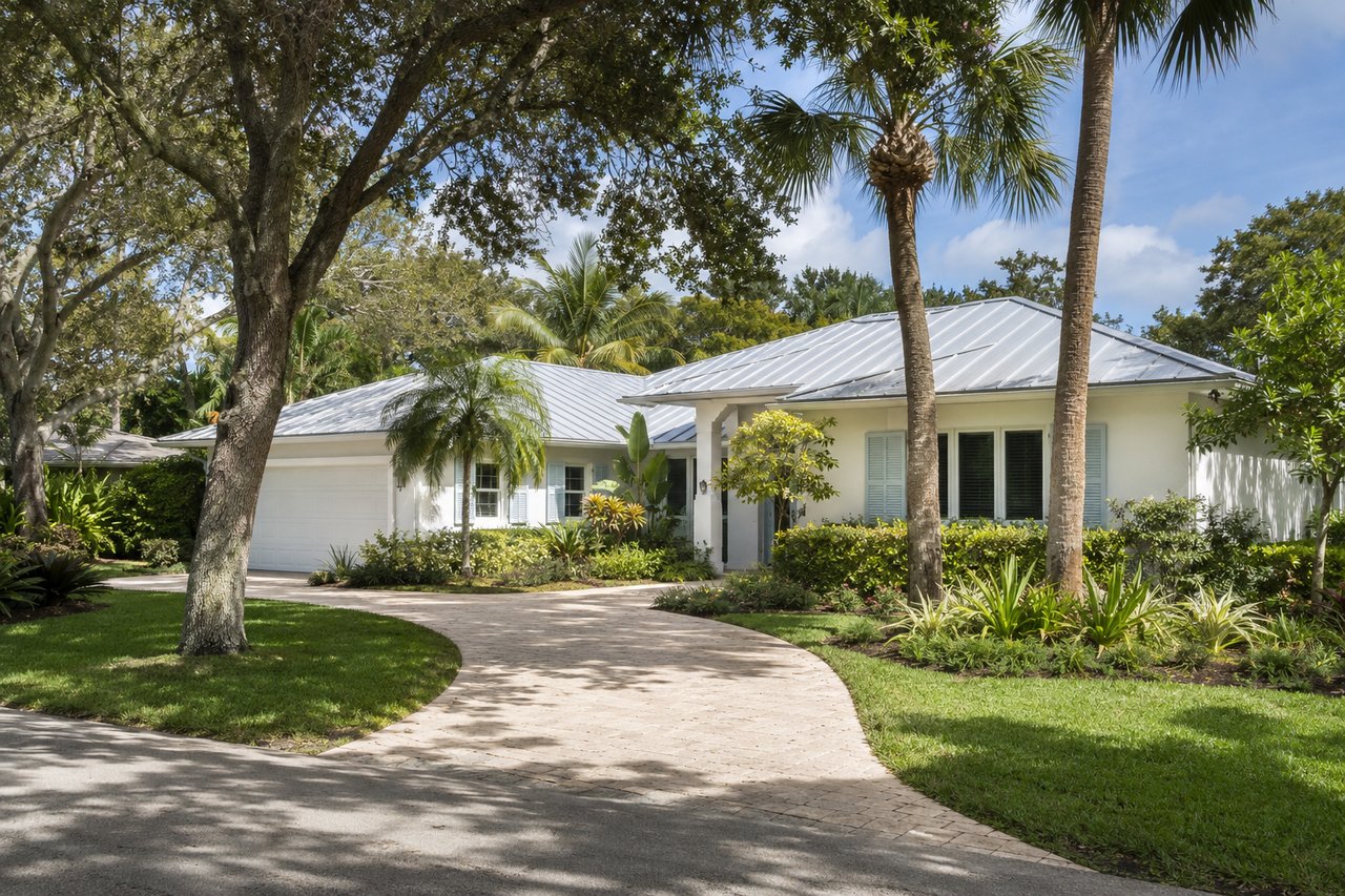 White single-story home with a metal roof, palms, and a curved paver driveway.