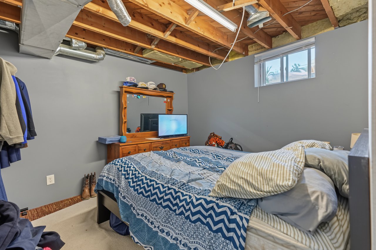 Bedroom with blue patterned bedding, wooden dresser, high window, and exposed ceiling joists.