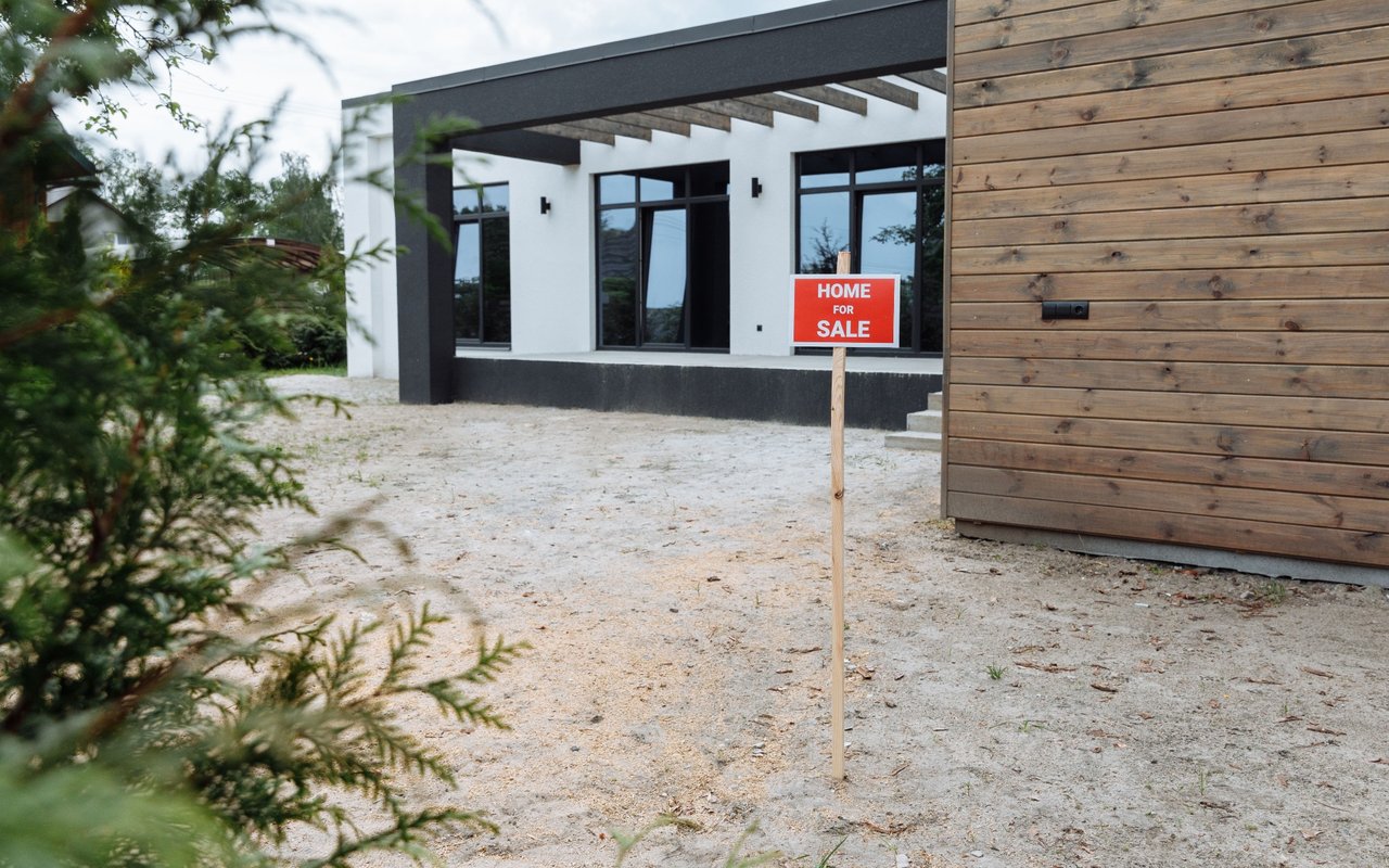 Modern home with a red for-sale sign in a gravel yard.