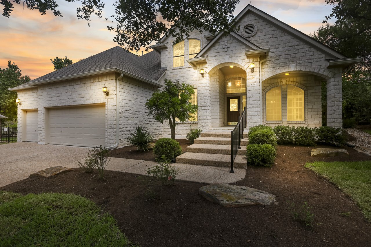 Two-story stone house with an arched entry, two-car garage, and illuminated windows at sunset.