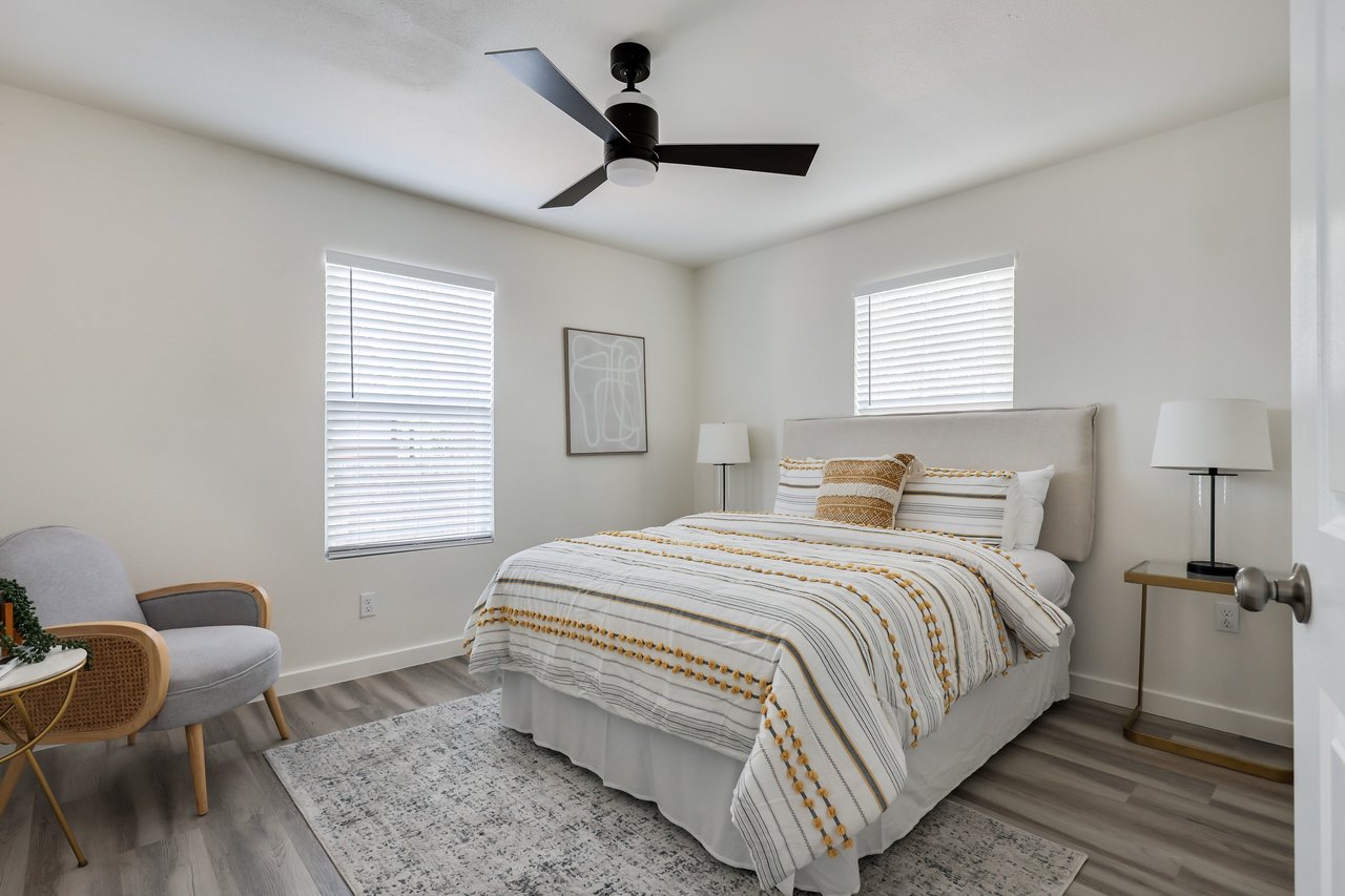 Furnished bedroom with striped white and mustard bedding, a black ceiling fan, and a gray chair.