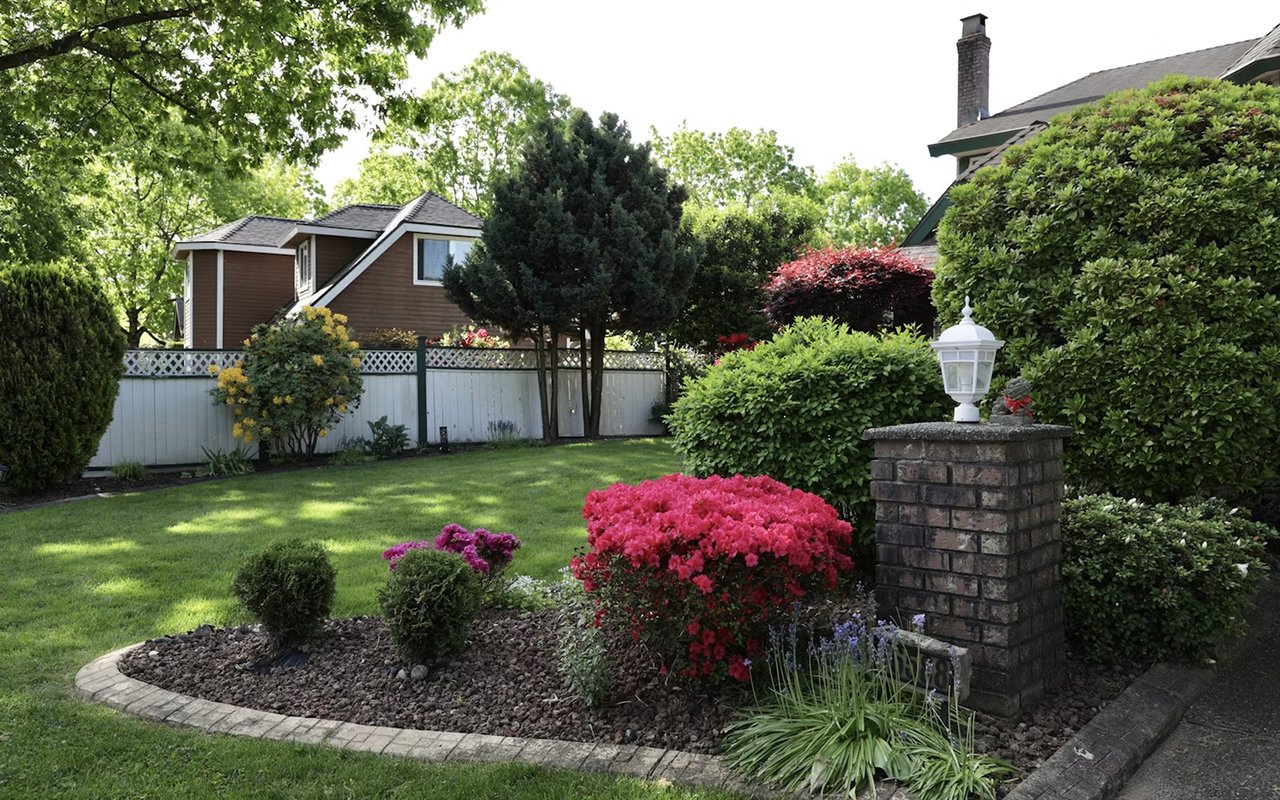 Landscaped yard with pink azaleas, a green lawn, mature trees, and a brick pillar with a lantern.