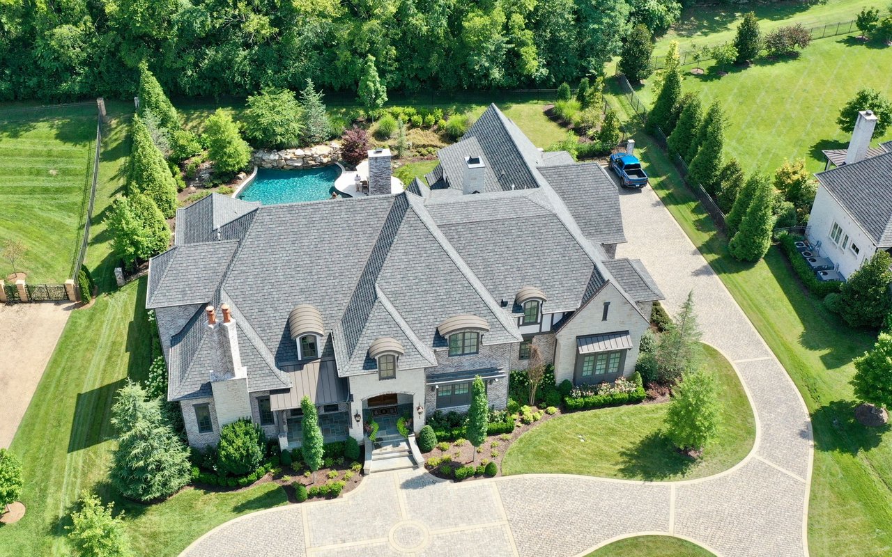 Aerial view of a stone home with a swimming pool and curved paver driveway.