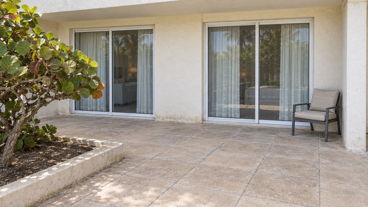 Pale stucco condominium facade with closed sliding-glass doors, a paved terrace, one outdoor chair, and a sea grape specimen in a raised concrete planter.