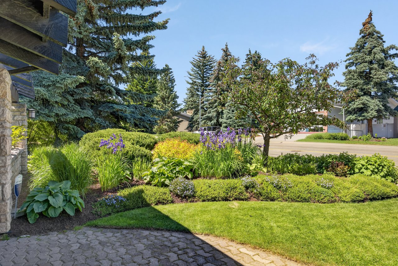 Landscaped front yard with a stone walkway, flowering beds, lawn, and mature evergreen trees.