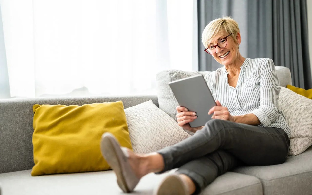 A smiling woman relaxes on a sofa while using a tablet.
