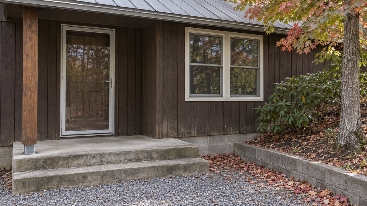 Dark wood cottage entry with paired windows, a gray metal porch roof, worn concrete steps and autumn leaves beside a gravel approach.