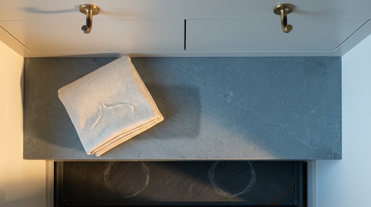 Soapstone entry bench with a folded cream throw, brass hooks, and a slate heated tray below