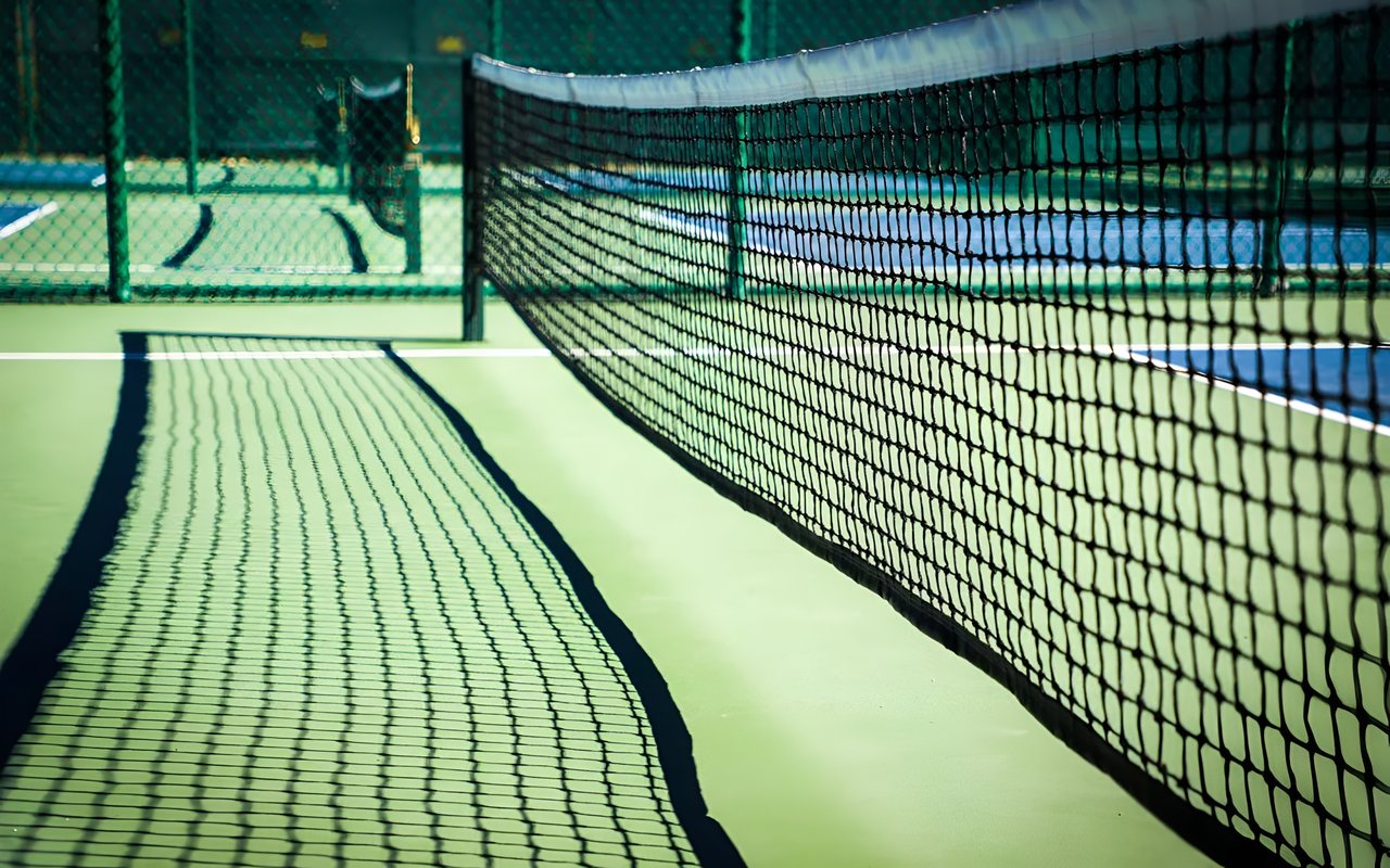 Close-up view of a tennis net casting a shadow on the green court with another court visible in the background.