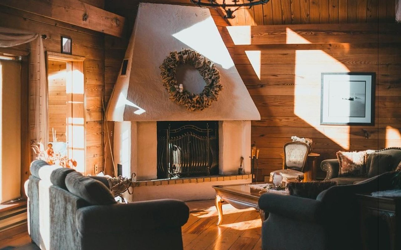 Rustic living room with a wreath above the fireplace, wood-paneled walls, and sunlight streaming in.