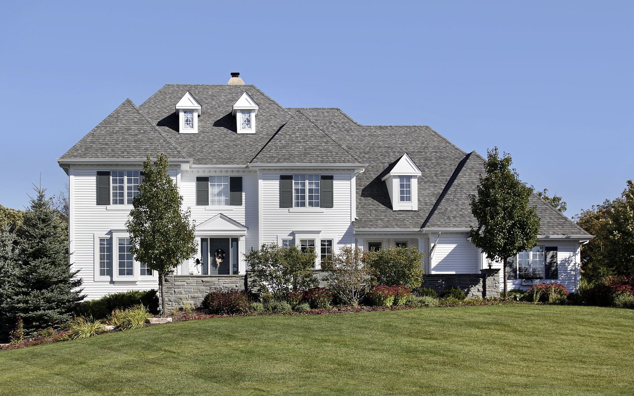 Large white house with gray roof, dormer windows, landscaped garden, and broad lawn.