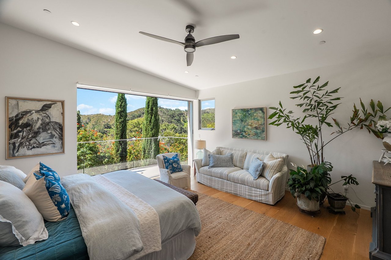 Furnished bedroom with a sitting area and wide glass doors overlooking green hills and cypress trees.