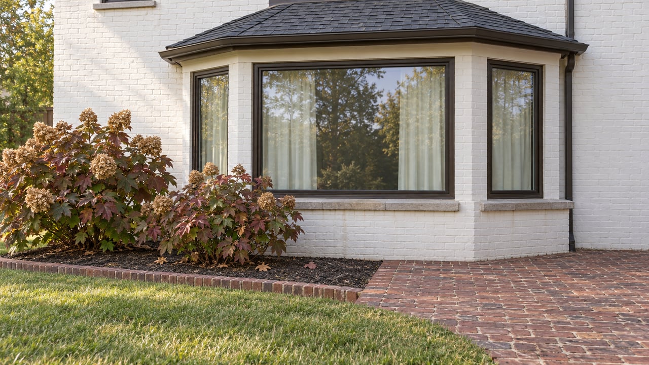 White-painted brick home with a dark-framed window bay above a weathered brick terrace, oakleaf hydrangeas and green lawn.