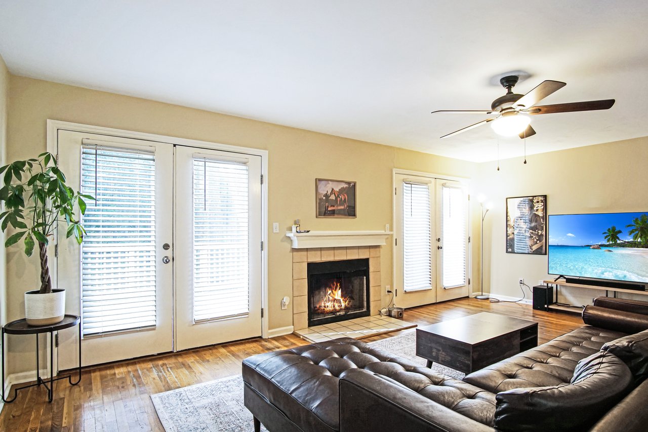 Living room at 529 Collinsford Road, Tallahassee, Florida, featuring a fireplace with tile surround, two sets of French doors with blinds leading to the back deck, and hardwood floors. The space includes a brown sectional sofa, coffee table, and ceiling fan, with natural light filling the room for a bright, welcoming atmosphere.