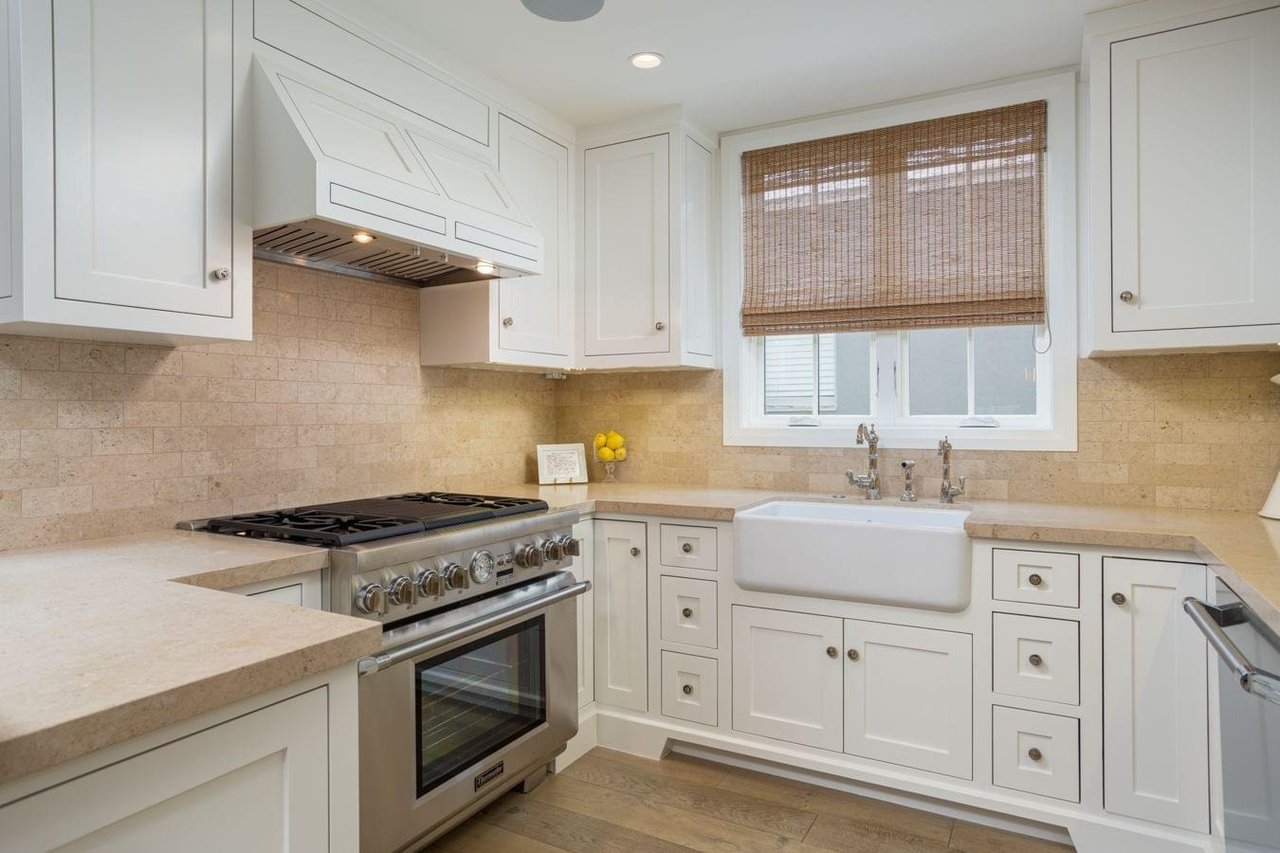  A bright kitchen with white cabinets, beige backsplash, a large farmhouse sink, and a professional-grade stainless steel range.