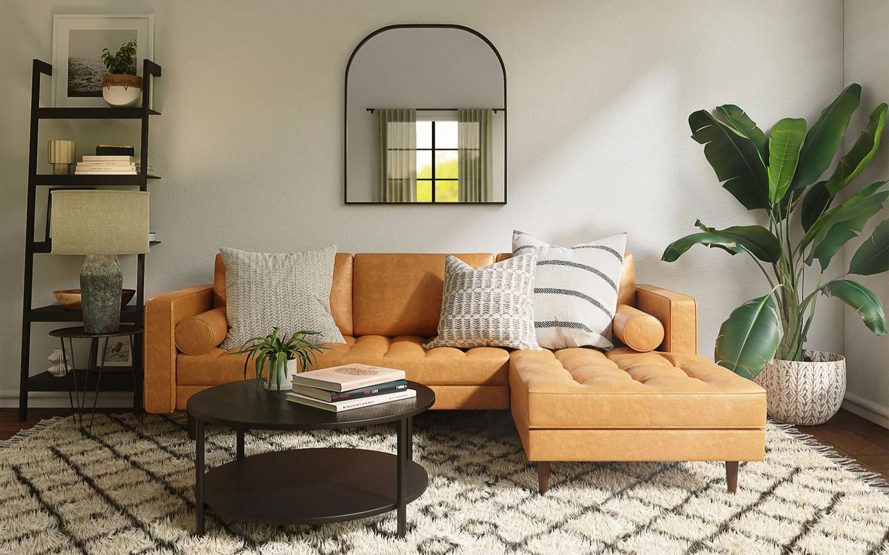 Staged living room with a caramel leather sectional, round coffee table, arched mirror, and leafy plants.