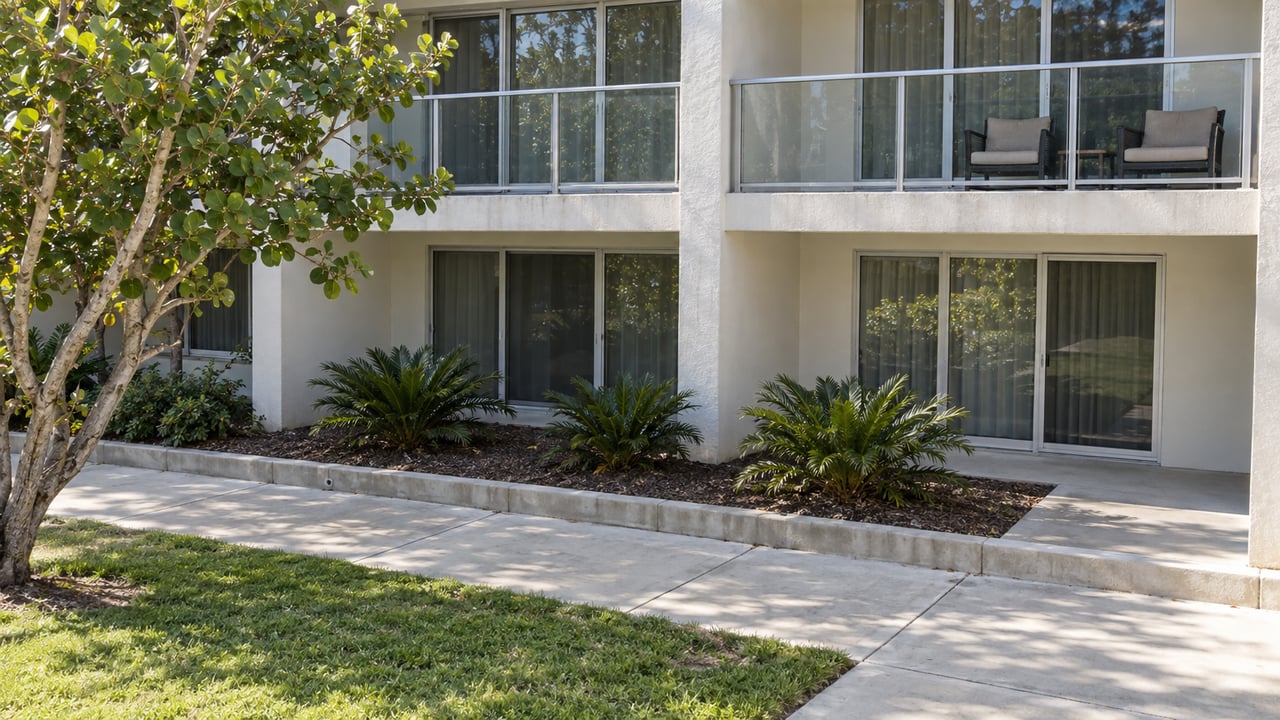 Sunlit condominium balconies overlook a modest shared concrete courtyard with a sea grape tree, coontie plants and a small lawn.